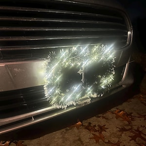 May include: A silver car's front grill with a wreath of white lights attached. The wreath is circular and has a frosted appearance. The lights are bright and illuminate the front of the car. Fallen leaves are on the ground.