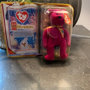 Puede incluir: Un oso de peluche Ty Beanie Baby rosa llamado Millennium, que lleva una cinta dorada y un amuleto de estrella dorada. El oso está empaquetado en una caja de plástico transparente con un fondo rojo, blanco y azul. El empaque presenta el logotipo de Ty y las palabras "Millennium the Bear".