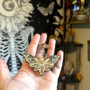 May include: A gold-colored metal keychain featuring a detailed moth design. The moth has intricate wing patterns and a skull-like head. The keychain is held up against a background with a skeleton and butterfly pattern.