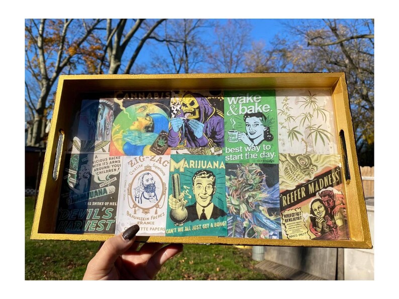 Puede incluir: Una bandeja rectangular de madera con un marco pintado de oro. El interior de la bandeja presenta un collage de im&aacute;genes y texto de estilo vintage relacionados con la cultura del cannabis, incluyendo las frases "Wake & Bake" y "Reefer Madness".