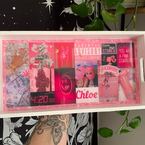 Puede incluir: Una bandeja rectangular blanca con un interior rosa que presenta varias im&aacute;genes rosas y rojas, incluyendo dinero, un esqueleto y el texto "PARENTAL ADVISORY". La bandeja tiene asas blancas y una capa protectora transparente.