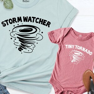 May include: A light blue t-shirt with the text "Storm Watcher" and a black tornado graphic. A pink baby onesie with the text "Tiny Tornado" and a white tornado graphic.