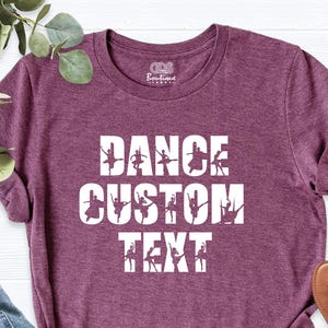 May include: A maroon t-shirt with the words "DANCE CUSTOM TEXT" in white block letters. The word "DANCE" is above the word "CUSTOM", and the word "TEXT" is below. Silhouettes of dancers are incorporated into the letters.
