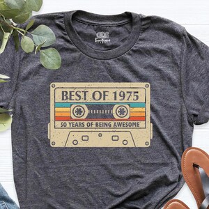 May include: Dark gray t-shirt featuring a retro cassette tape design with the text "BEST OF 1975" and "50 YEARS OF BEING AWESOME". The shirt is paired with blue jeans and brown sandals.