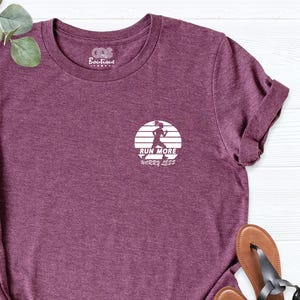 May include: A maroon t-shirt with a small white graphic of a runner inside a circle, with the words "Run More Worry Less". The shirt is paired with blue jeans and brown sandals.