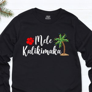 May include: Black long-sleeve shirt with a white hibiscus flower, palm tree with Christmas lights, and the text "Mele Kalikimaka".