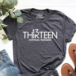 May include: A dark gray t-shirt with white text that reads "13 THIRTEEN #OFFICIAL TEENAGER". The shirt is laid out on a white wooden surface with a pair of brown sandals and blue jeans.