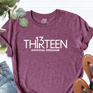 May include: A maroon t-shirt with the text "13 THIRTEEN #OFFICIAL TEENAGER" printed in white.