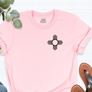 K&ouml;nnte beinhalten: Rosa T-Shirt mit einem schwarzen Zia-Symbol auf der Vorderseite. Das T-Shirt ist aus einem weichen, bequemen Material gefertigt und hat eine lockere Passform.