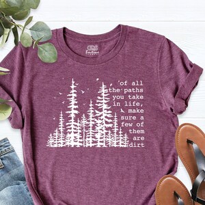 Op de afbeelding: Een maroon t-shirt met een witte afbeelding van een bos en de tekst 'of all the paths you take in life, make sure a few of them are dirt'.