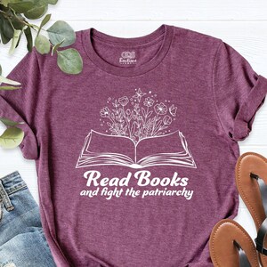 May include: A maroon t-shirt with a white graphic of an open book with flowers growing out of it. The text "Read Books and fight the patriarchy" is printed below the book.