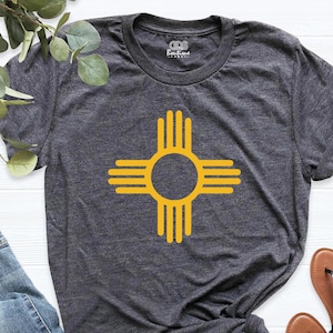 May include: Dark gray t-shirt featuring a yellow Zia symbol, a Native American symbol from New Mexico. The shirt is made of soft material and has a crew neck. The Zia symbol is centered on the front of the shirt. The shirt is paired with blue jeans and sandals.