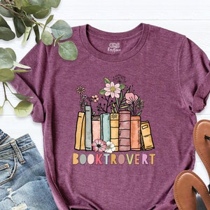 May include: A purple t-shirt featuring a graphic of a stack of colourful books with flowers on top. The text "BOOKTROVERT" is printed below the books.