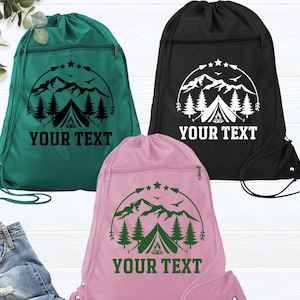 May include: Three drawstring backpacks in teal, black, and pink. Each bag features a mountain and tent graphic with the text "YOUR TEXT" in a semi-circle. The bags have a zippered pocket and a drawstring closure.