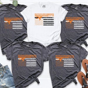 May include: A collection of t-shirts in various shades of gray and white, each featuring a design related to Multiple Sclerosis Awareness. The design includes an orange ribbon, stars, and stripes, along with the text "Multiple Sclerosis Awareness John Team."