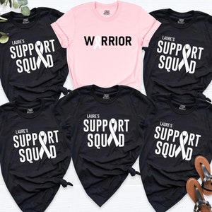 May include: Five black t-shirts with white text and a white ribbon design. The text on the shirts reads "Laure's Support Squad". One pink t-shirt with the text "Warrior" and a white ribbon design.