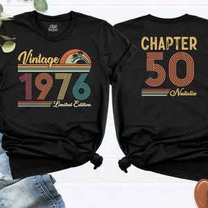 May include: Black t-shirts with retro-style graphics. One shirt features "Vintage 1976" with a sunset design, the other says "Chapter 50 Natalie". Both shirts are short-sleeved and have a crew neck.