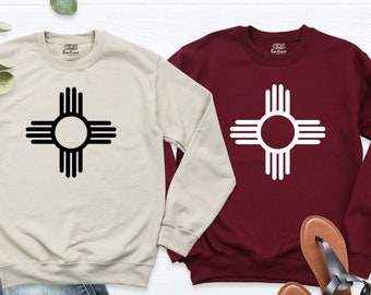 Könnte beinhalten: Zwei langärmelige Sweatshirts mit einem schwarzen und weißen Zia-Symbol auf beigefarbenem und maroonfarbenem Hintergrund. Die Sweatshirts liegen auf einer weißen Oberfläche mit braunen und schwarzen Sandalen.