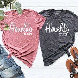 May include: Two t-shirts, one pink and one grey, with the text "Abuelita Est. 2021" and "Abuelito Est. 2021" in white.
