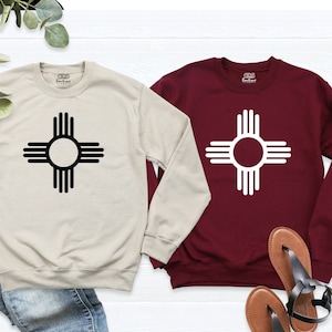 May include: Two long-sleeved sweatshirts with a black and white Zia symbol on a beige and maroon background. The sweatshirts are laid out on a white surface with brown and black sandals.
