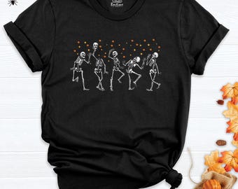 May include: A black short-sleeved t-shirt featuring a graphic print of five white skeletons dancing. The skeletons are surrounded by small orange pumpkins and yellow stars. The inner neck label displays the text CDS Boutique.