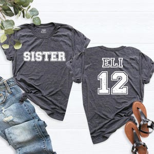 May include: Two heather grey t-shirts. One shirt says "Sister" in white block letters. The other shirt says "Eli 12" in white block letters with a distressed look.