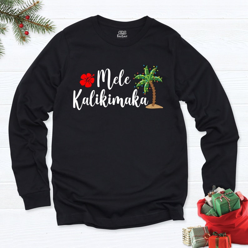 May include: Black long-sleeve shirt with a white hibiscus flower, palm tree with Christmas lights, and the text "Mele Kalikimaka".