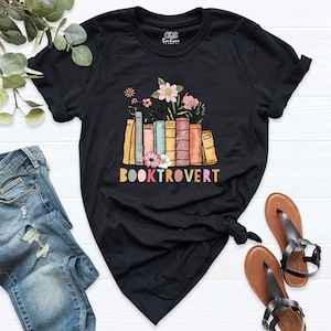 May include: Black t-shirt featuring a graphic of a stack of colourful books with flowers on top. The text "BOOKTROVERT" is printed below the books in rainbow colours.
