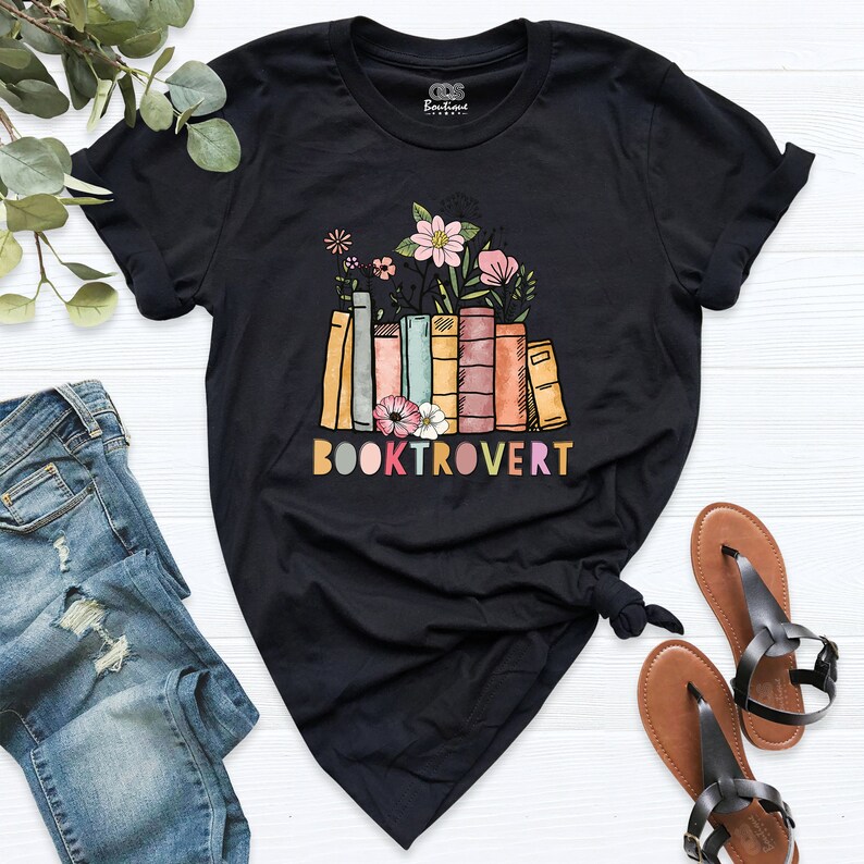 May include: Black t-shirt featuring a graphic of a stack of colourful books with flowers on top. The text "BOOKTROVERT" is printed below the books in rainbow colours.