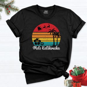 Może przedstawiać: Czarny t-shirt z krótkim rękawem z grafiką zachodzącego słońca w poziome paski w kolorach czerwonym, pomarańczowym, żółtym i niebieskim. Wzór zawiera sylwetkę Świętego Mikołaja w saniach, palmy oraz kwiat hibiskusa, z napisem Mele Kalikimaka.