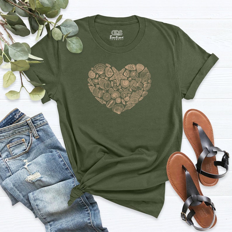 May include: A green t-shirt with a heart-shaped design made of seashells. The design is in a light brown color.