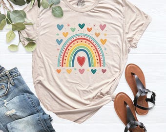 May include: A beige short-sleeve t-shirt featuring a central graphic of a colorful rainbow surrounded by small hearts and stars. The rainbow consists of red, orange, yellow, green, and blue stripes. A red heart is positioned below the rainbow.