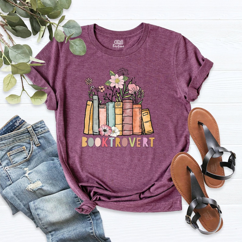 May include: A purple t-shirt featuring a graphic of a stack of colourful books with flowers on top. The text "BOOKTROVERT" is printed below the books.