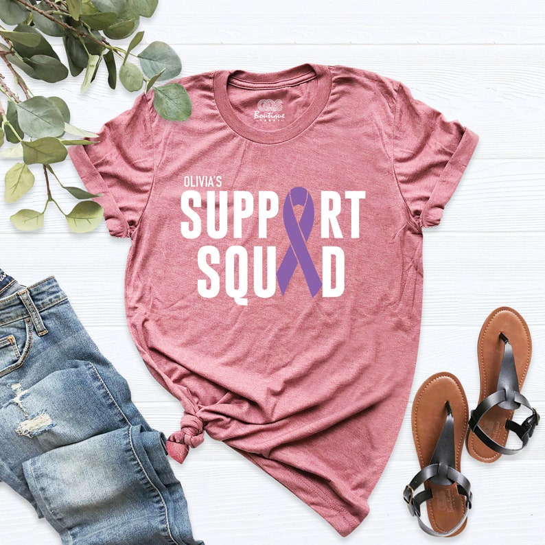 K&ouml;nnte beinhalten: Ein mauvefarbenes T-Shirt mit dem Text "Olivia's Support Squad" in Wei&szlig; und einem lilafarbenen Band.