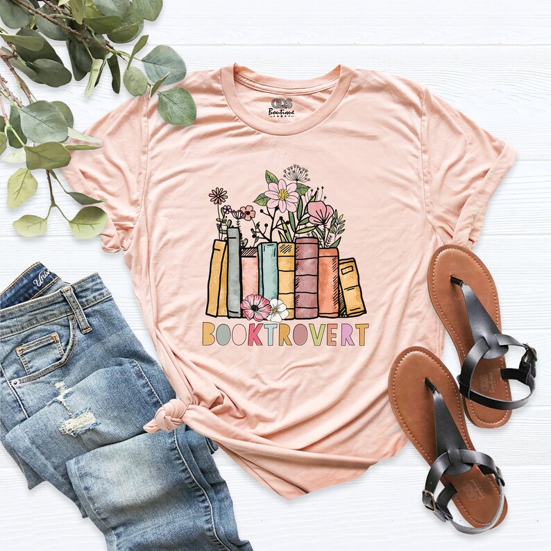 May include: A light pink t-shirt featuring a graphic of a stack of colourful books with flowers on top. The text "BOOKTROVERT" is printed below the books.