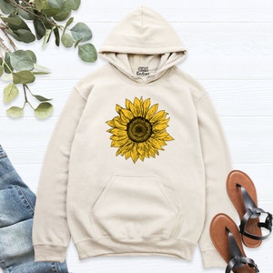May include: A cream-colored hoodie with a sunflower graphic printed on the front. The hoodie has a drawstring hood and a kangaroo pocket.