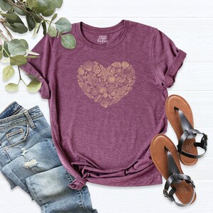 May include: A maroon t-shirt with a gold heart-shaped design made of seashells and starfish. The shirt is made of a soft, comfortable material.