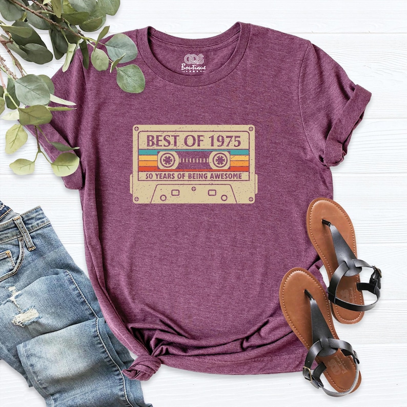 May include: A maroon t-shirt featuring a retro cassette tape design with the text "BEST OF 1975" and "50 YEARS OF BEING AWESOME". The shirt is paired with blue jeans and brown sandals.