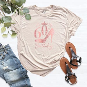 May include: A light beige t-shirt with a rose gold graphic that says "It's My 60th Birthday" with a crown and a high heel shoe made of dots.