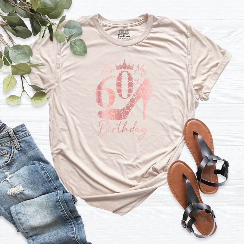 May include: A light beige t-shirt with a rose gold graphic that says "It's My 60th Birthday" with a crown and a high heel shoe made of dots.