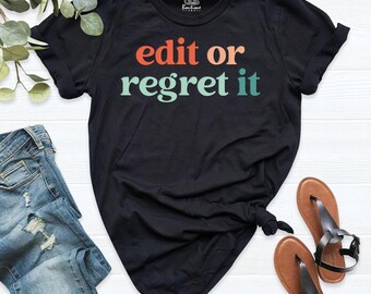 Op de afbeelding: Een zwart t-shirt met korte mouwen en de tekst edit or regret it in oranje en lichtgroene letters. Het shirt heeft een ronde hals en een klein merklogo aan de binnenkant van de kraag. Een blauwe spijkerbroek en bruine sandalen liggen ernaast.