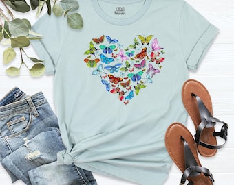 May include: A light blue short-sleeve t-shirt featuring a heart-shaped graphic composed of various colorful butterflies. The inner neck label displays the text ODS Boutique. The shirt is paired with blue denim jeans and brown sandals.