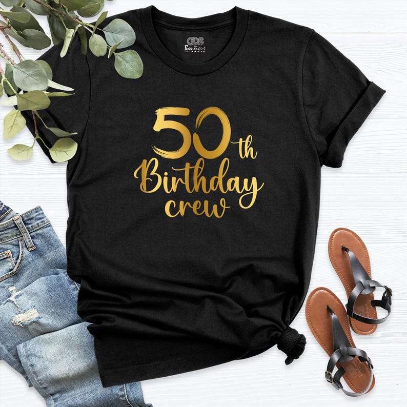 May include: Black crew neck t-shirt with gold text that reads "50th Birthday crew". The shirt is styled with rolled-up sleeves and is paired with blue jeans and brown sandals. The shirt is a casual wear item.