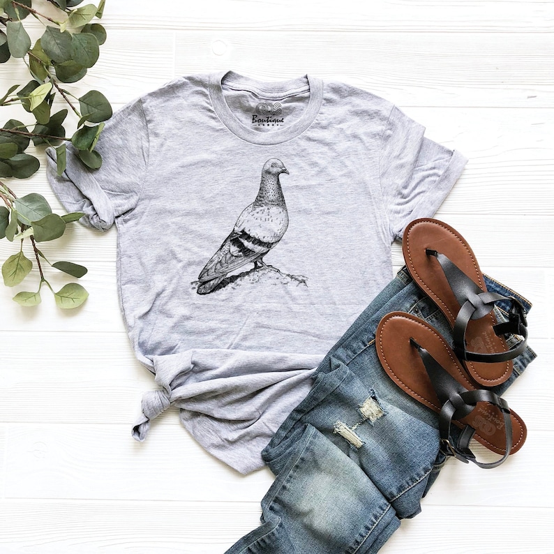 Op de afbeelding: Een heidekleurig T-shirt met een zwart-witte illustratie van een duif. Het T-shirt heeft een ronde hals en korte mouwen. Het T-shirt ligt plat op een wit oppervlak met een paar bruine leren sandalen en een blauwe jeans.