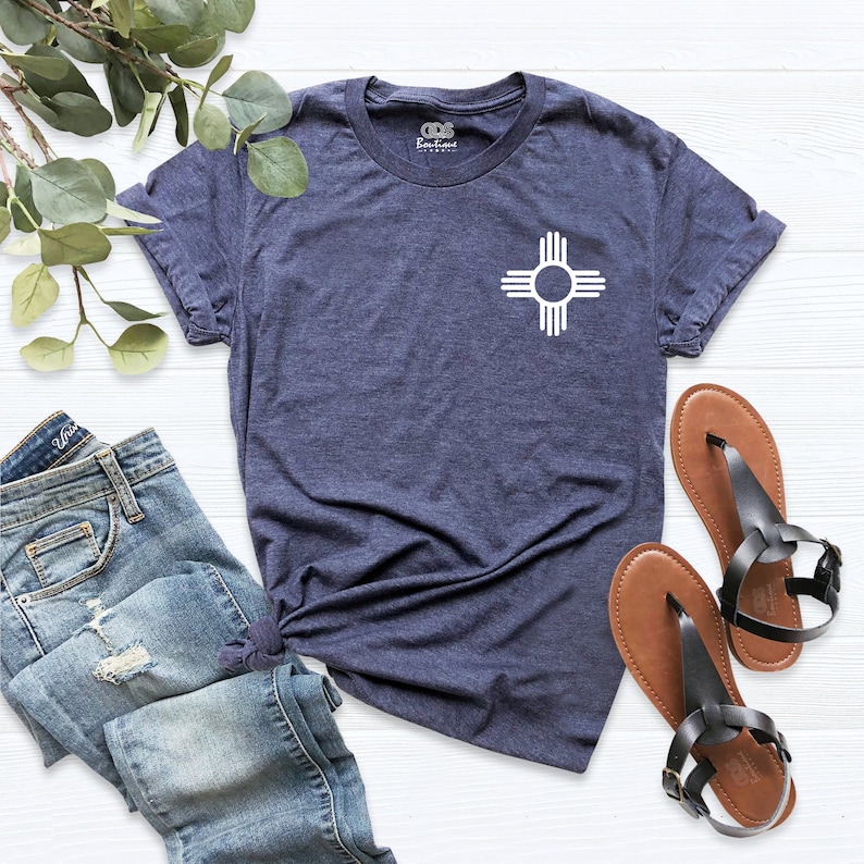 K&ouml;nnte beinhalten: Ein marineblaues T-Shirt mit einem wei&szlig;en Zia-Symbol auf der linken Brust. Das T-Shirt liegt flach auf einer wei&szlig;en Holzoberfl&auml;che mit einem Paar braunen Sandalen und einer blauen Jeans.