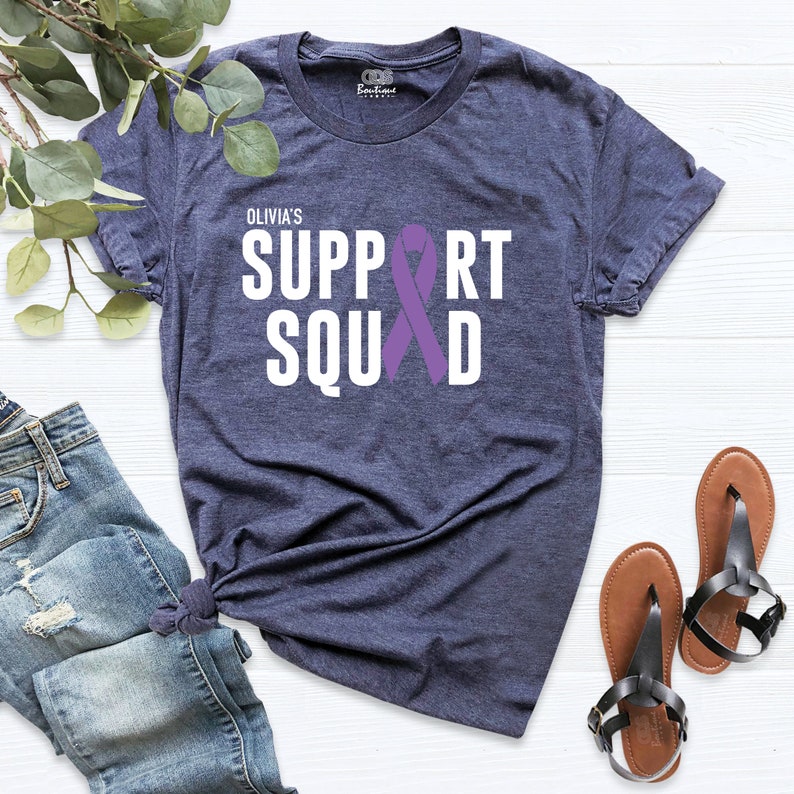 K&ouml;nnte beinhalten: Ein marineblaues T-Shirt mit dem Text "Olivia's Support Squad" in wei&szlig;en Buchstaben und einem lila Band.