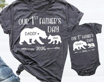 May include: A dark heather gray t-shirt and a matching infant bodysuit, both featuring white screen-printed graphics of a large bear labeled Daddy and a small bear labeled Jack, with the text Our 1st Father's Day and the year 2026.