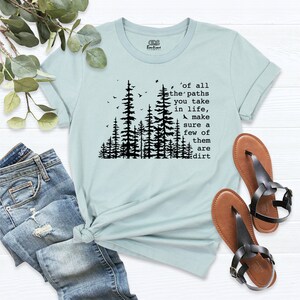 Op de afbeelding: Lichtblauw T-shirt met een zwart silhouet van een bos en de tekst 'of all the paths you take in life, make sure a few of them are dirt'.