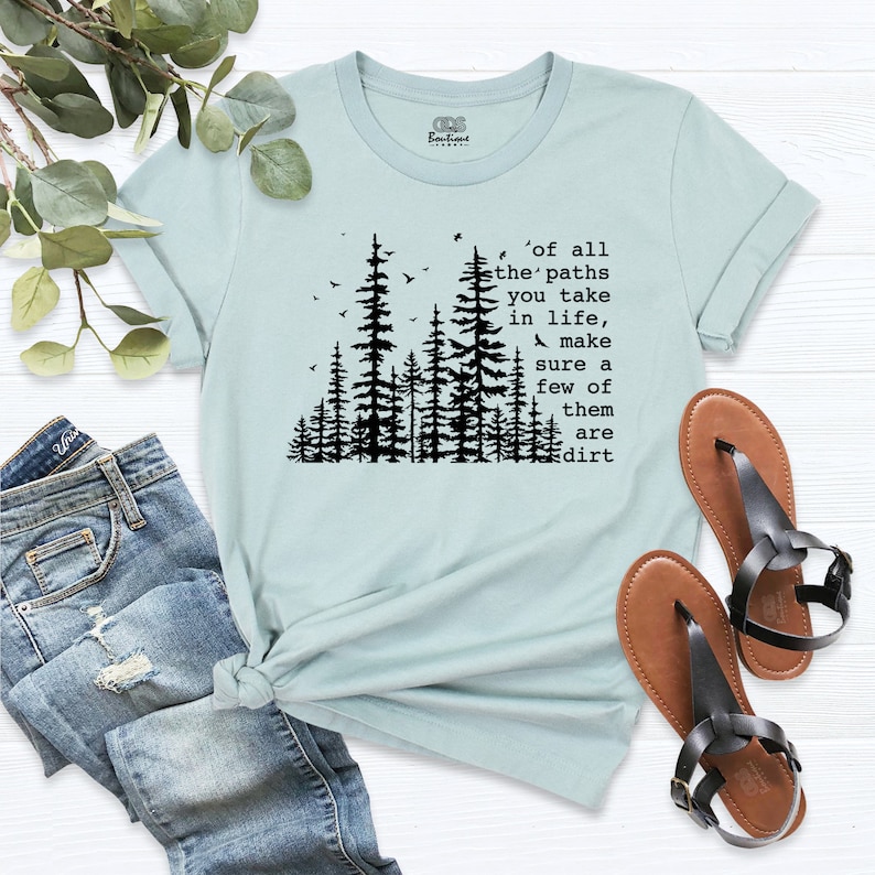 Op de afbeelding: Lichtblauw T-shirt met een zwart silhouet van een bos en de tekst 'of all the paths you take in life, make sure a few of them are dirt'.