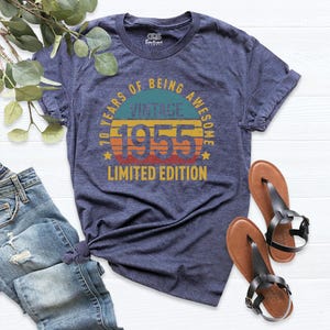 Op de afbeelding: Een marineblauw T-shirt met een vintage ontwerp uit 1955. Het shirt heeft een zonsondergangafbeelding met de tekst "70 Years of Being Awesome Vintage 1955 Limited Edition".